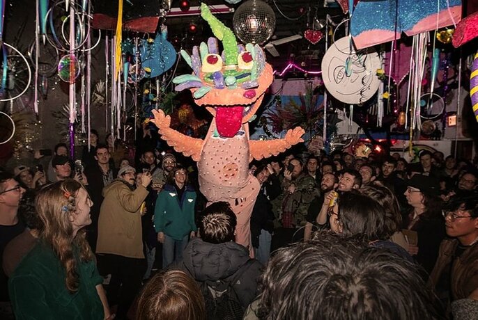 Night time and outdoors with a large crowd of people gathered around a giant walking puppet, with a fantastical head; in the background are art installations and hanging decorations.