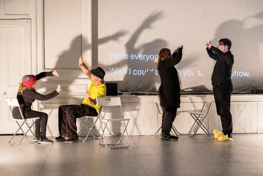 Four people in an open space performing movements together, a light casting their shadows on the wall. 