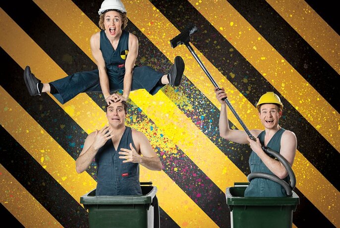 A woman leapfrogs over a man standing in a wheelie bin, next to another man in a wheelie bin holding a vacuum cleaner. All are in construction outfits and hard hats.