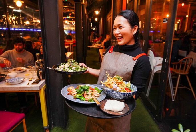Waiter carrying tray of food in left hand and dish in right, looking away to left and smiling, nicely lit restaurant behind and to left.