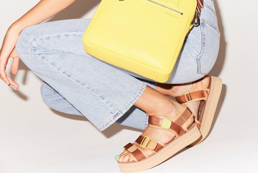 A model crouching down in a pair of bronze and lime green chunky and strappy platform sandals.