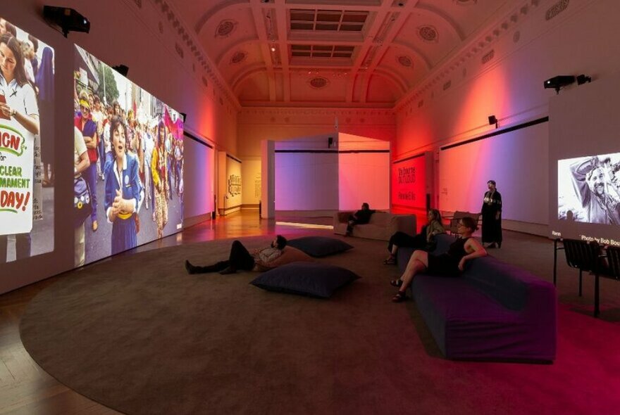 Installation view of the Rennie Ellis exhibition in a dimly lit room at the State Library, showing large photographs projected onto the walls, with people reclining on bean bags and lounges viewing them.