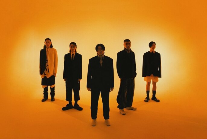 Five members of the band Sunset Rollercoaster lined up in a V formation, all wearing dark clothing and standing against an orange backdrop.