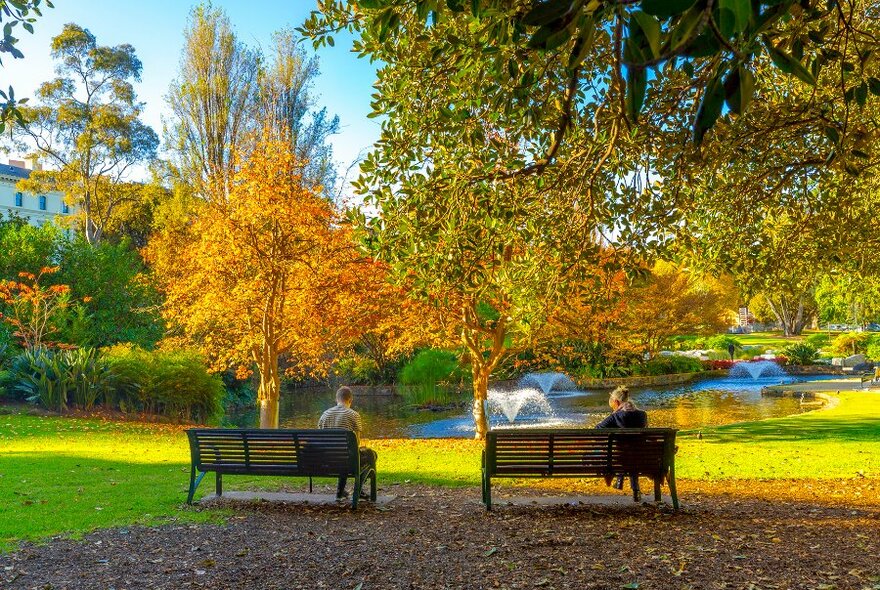 Two people sitting on two benches in a park by a lake.