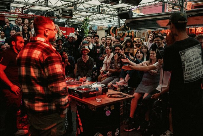 Crowd of onlookers in an outdoor club or bar, watching a person rapping into a microphone in front of a DJ mixing desk of turntables.