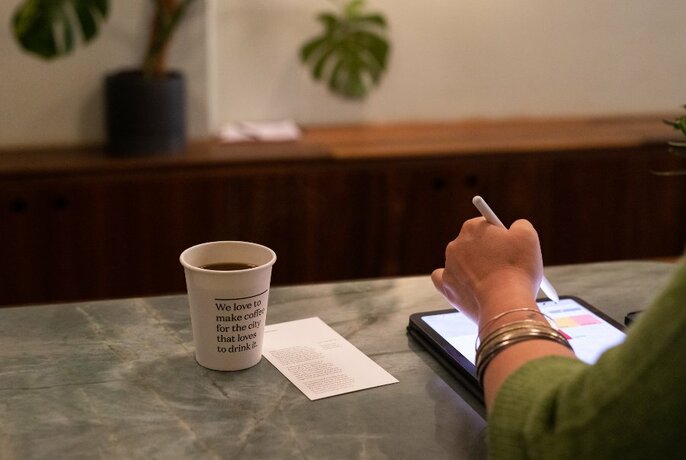 Person seated at a table with a cup of coffee next to them as they read from a digital tablet.
