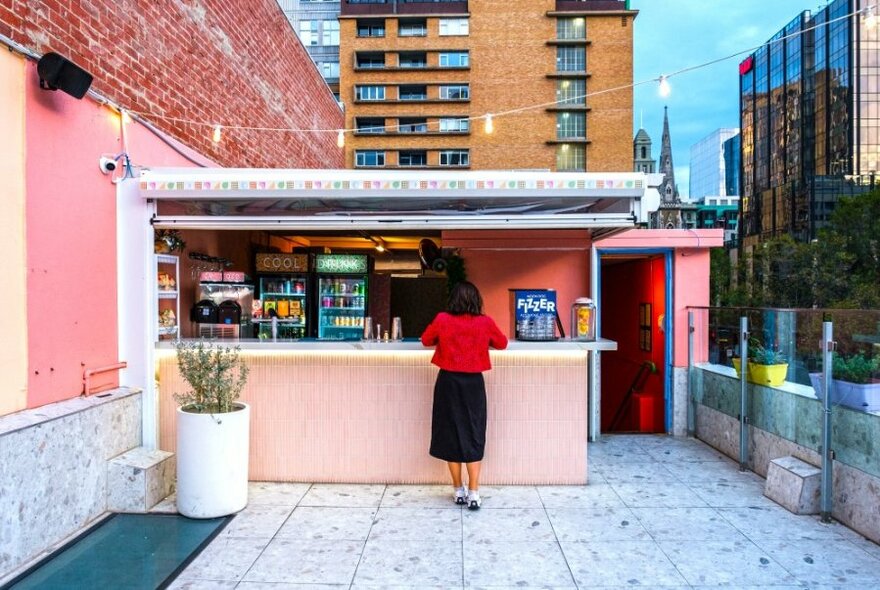 A woman standing at a pastel pink bar on a city rooftop.