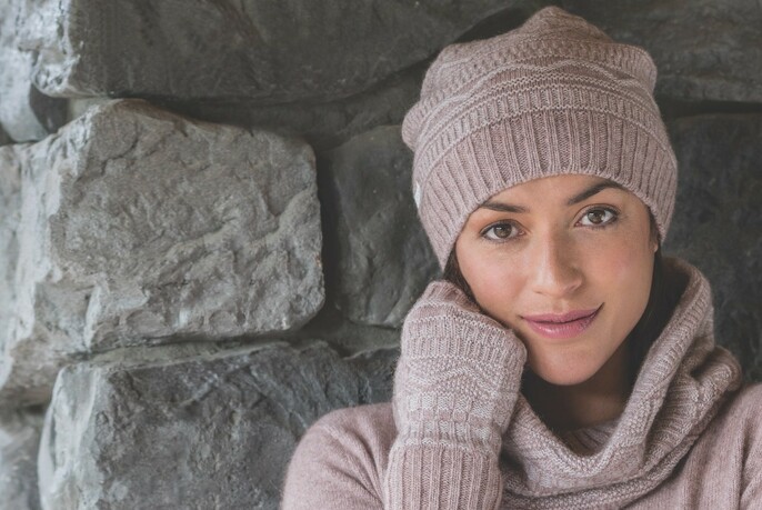 Model wearing lilac knitted hat, gloves, scarf and jumper, standing in front of rough-hewn rock wall.