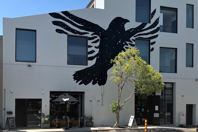 Cafe exterior with huge mural of a black bird, outside tables with umbrella beside tree.