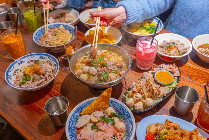 Someone reaching for a noodle dish with chopsticks. The table is set with lots of colourful Thai dishes.