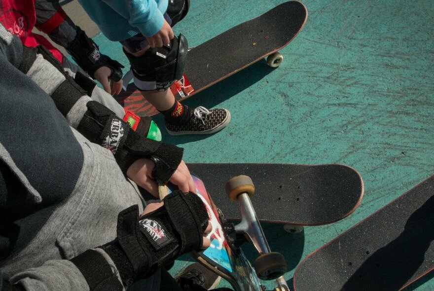 The legs and torsos of children wearing protective skate gloves and knee pads, holding their skateboards and waiting for their turn to skate down a ramp.