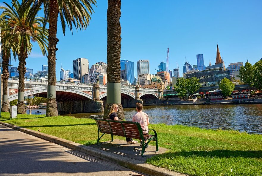 Two people sitting on a bench beside a city river on a sunny day. 