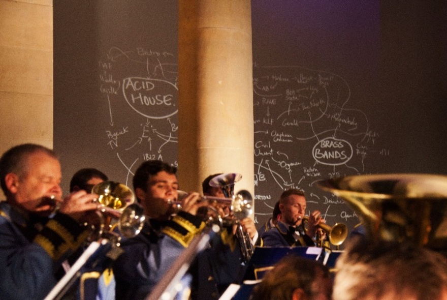A brass band performing inside a dimly lit space, a wall behind the musicians decorated with a chalk drawn mind-map.