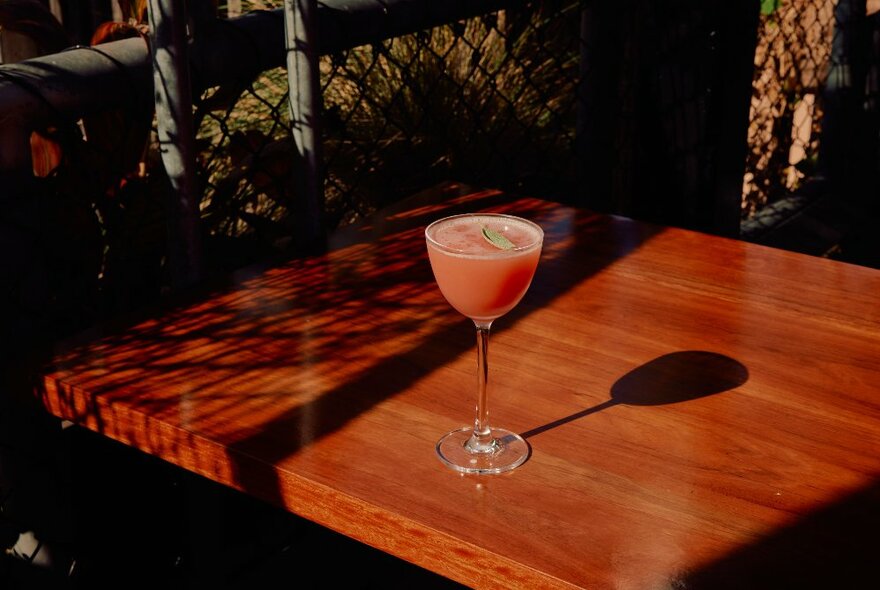 A pink coloured cocktail on an outdoor wooden table, in the sunlight and shadows.