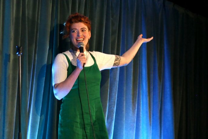 Woman in white t-shirt under green, scrappy dress; smiling, holding mic with right arm and gesturing up with left arm, in front of a blue curtain.