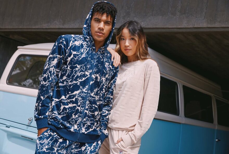 Man and woman posing in Fila leisure tracksuits in front of a pale blue Kombi van.