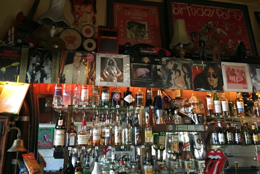 Crowded hotel bar with rows of bottles and rock posters with Sid Vicious, Robert Plant and Rolling Stones tongue.