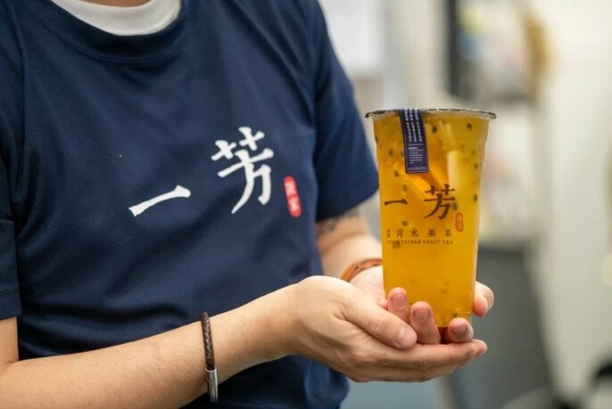 Hands holding a single tall sealed plastic cup of  fruit tea in front of their torso.