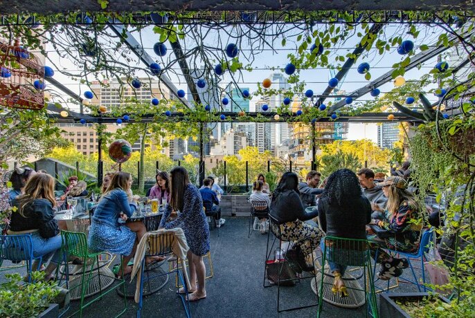 A busy rooftop bar in the day with patrons at tables and vines and lanterns hanging above.
