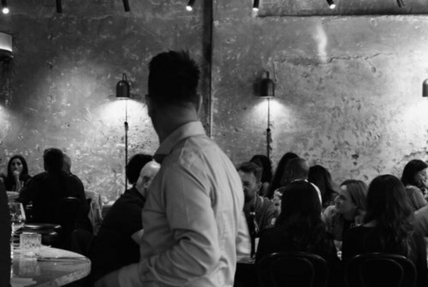 Waiter turning towards packed tables of diners in a restaurant with moody downlighting.