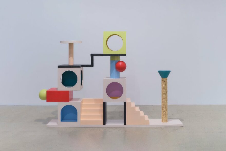 A large coloured sculpture in a large empty room, comprising small steps, cubes and scratch posts, all joined together, for a cat to play on.
