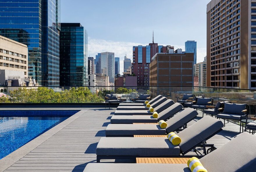 Rooftop pool deck at Le Méridien Melbourne with lounge deck chairs, a pool, plants and a city skyline view. 