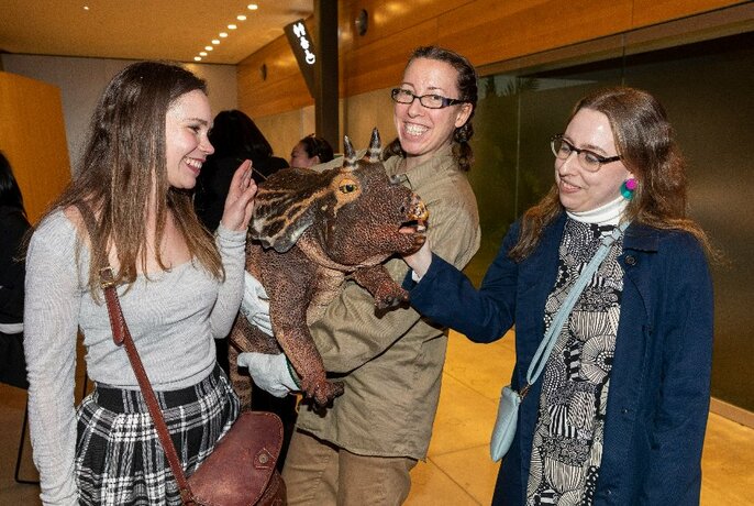 Three women standing smiling while patting a model dinosaur.