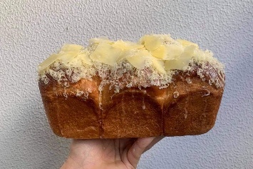 A brioche-type bread topped with cheese and held up against a white wall by a single hand.
