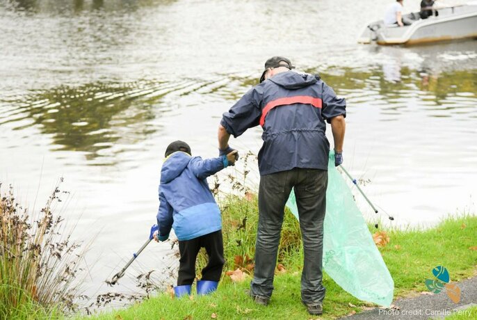 Child and adult at river's edge, seen from behind, with long rubbish tongs and plastic bag.
