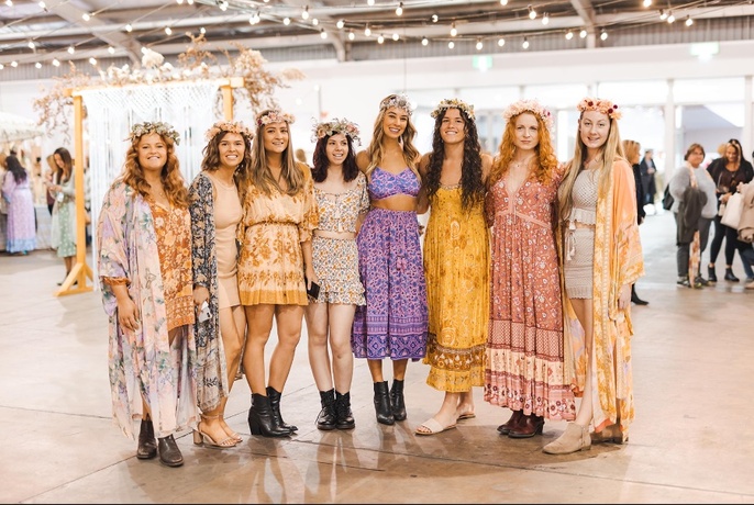 Row of women in flower-print clothes, and head garlands, inside an exhibition hall.