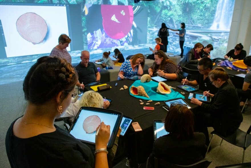 A group of people participating in a drawing workshop, some seated on couches, others at a table, drawing on ipads, while large images of objects are projected on the walls around them.