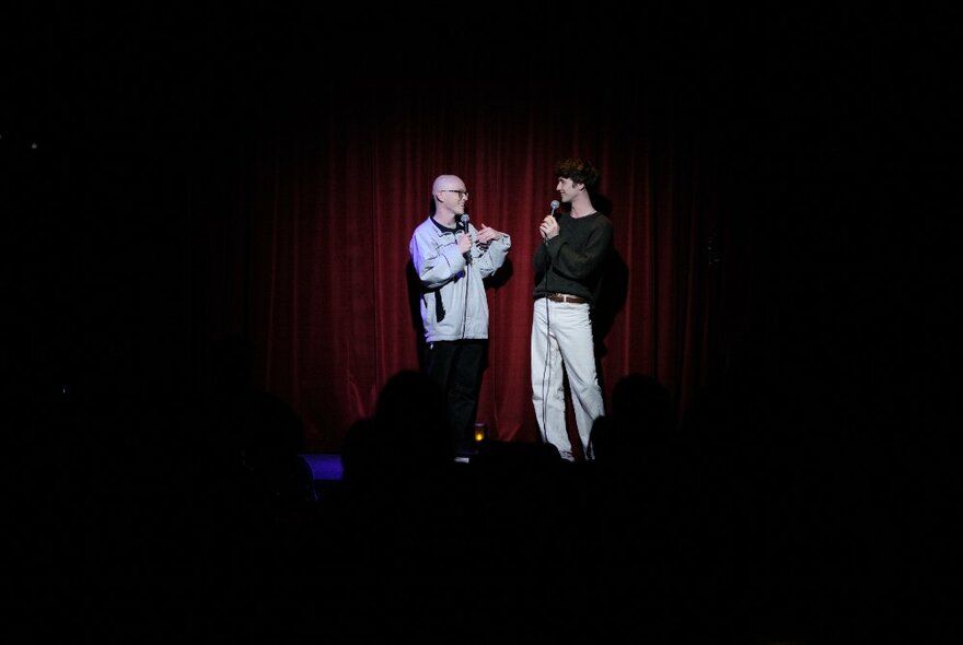 Two stand-up comedians on a darkened stage, each holding microphones.