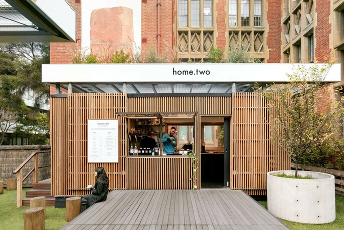 Outdoor cafe with slatted panelling in Melbourne University courtyard with person seated on a wooden platform, barista at the counter.