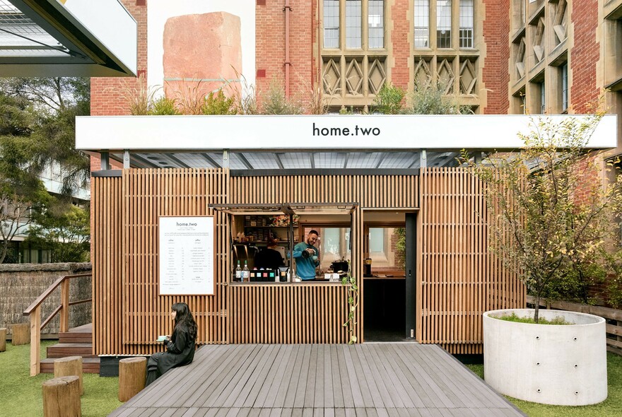 Outdoor cafe with slatted panelling in Melbourne University courtyard with person seated on a wooden platform, barista at the counter.