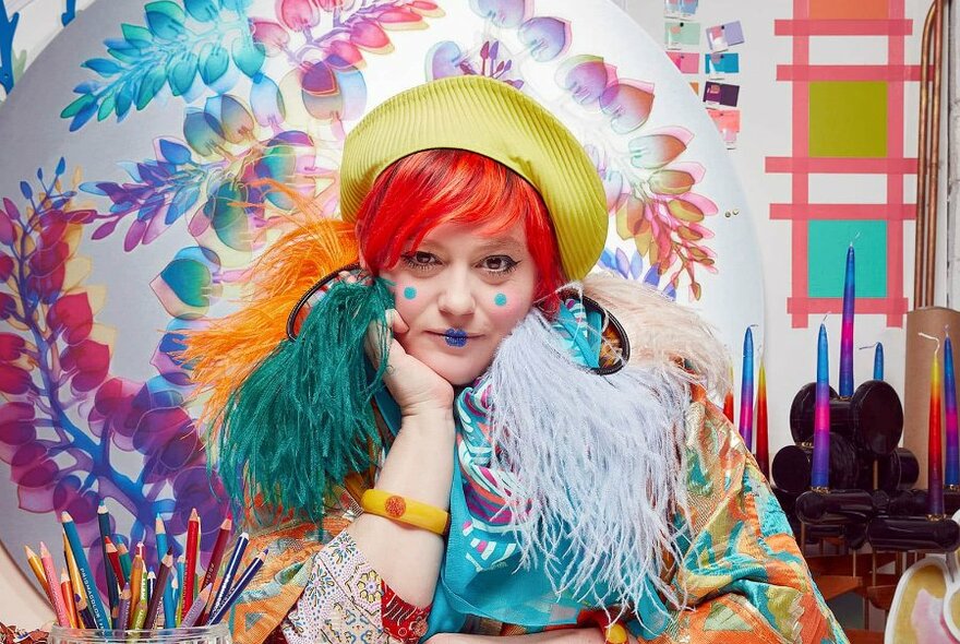 Photo portrait of designer Bethan Laura Wood wearing brightly coloured garments, a yellow hat, feathers around her neck, her chin resting on her hand, posing in a decorative space, a container of coloured pencils on the desk in front of her.