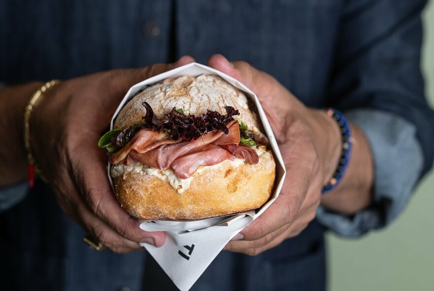 A wrapped cured meat panini, being held by two hands. 