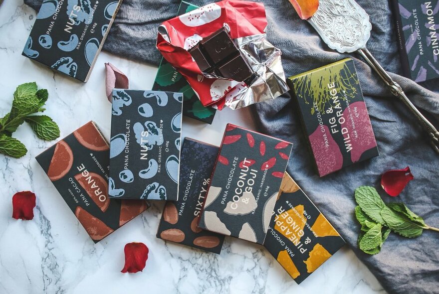 A selection of chocolate bars with coloured labels scattered on a surface of fabric and rose petals