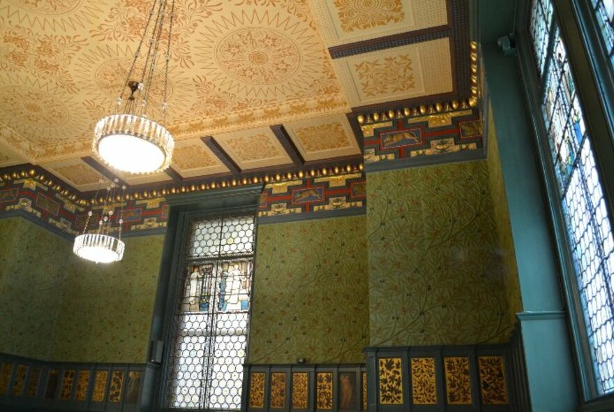 The interior of a period room with ornate patterned wallpaper on the walls and ceiling, wainscoting and stained glass windows.
