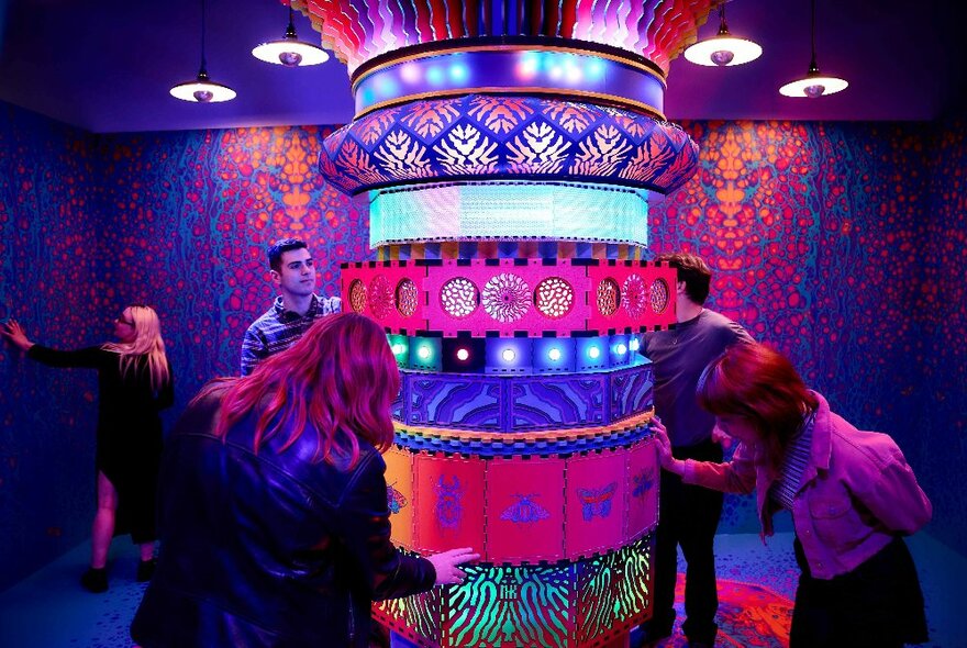People interacting with a colourful, cylindrical artistic installation with neon lights and varied textures.