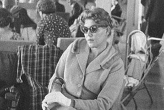 Black and white photo of a woman wearing sunglasses, gloves and a coat, seated in a chair, with people in the background, circa mid-1950s.