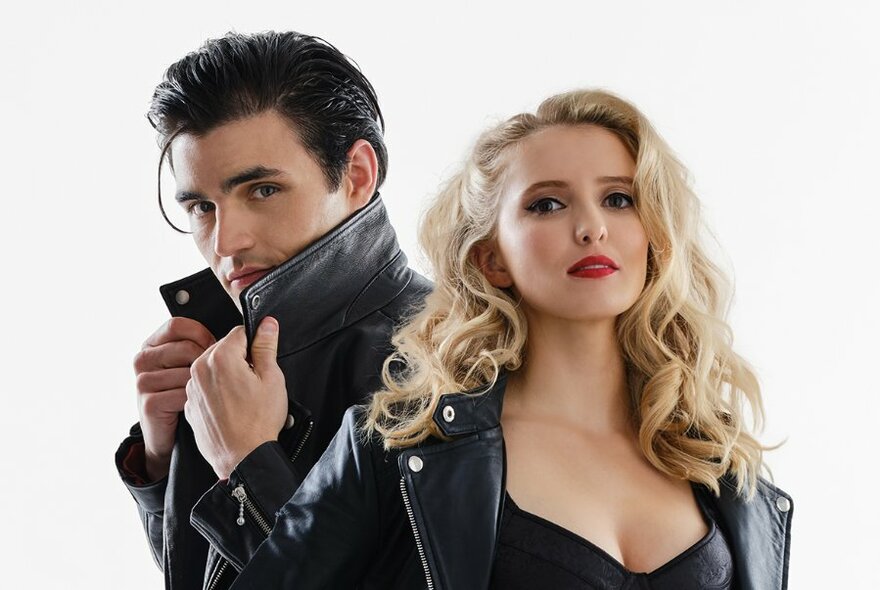 Two actors dressed as the characters Danny and Sandy from the musical Grease - Danny holding up the collar of his black leather jacket behind Sandy with curled long blonde hair, also wearing a leather jacket. 