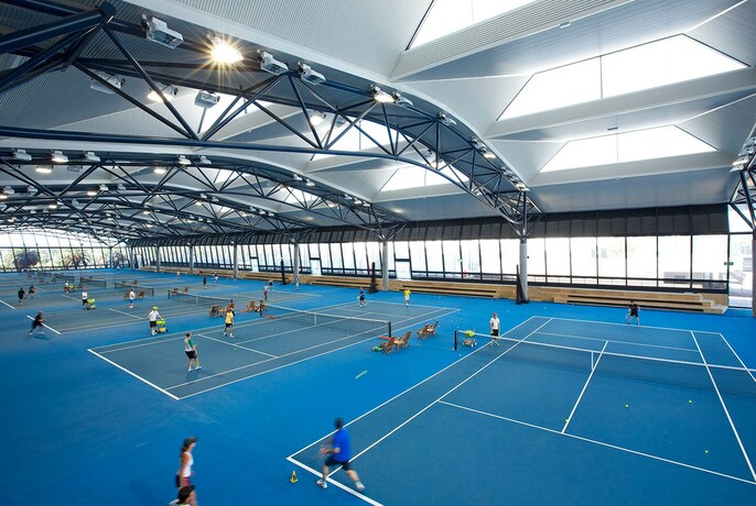 Large indoor area with several tennis courts with people playing on a bright blue surface.