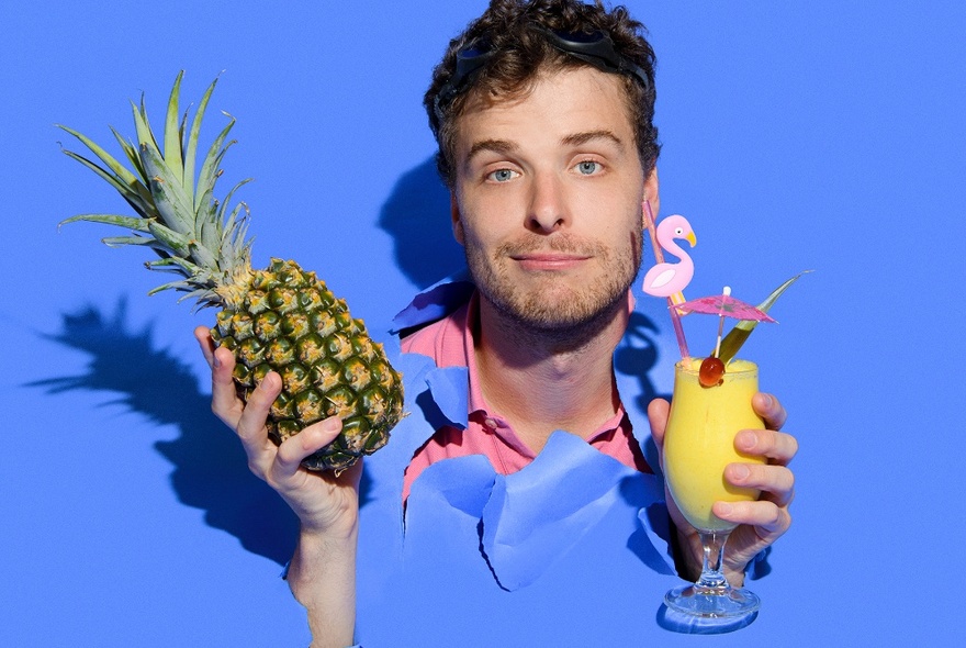 Man's head and hands poking out of torn rips in blue paper background screen, with a wry smile on his face, holding a whole pineapple in one hand and a pina colado cocktail in the other hand.