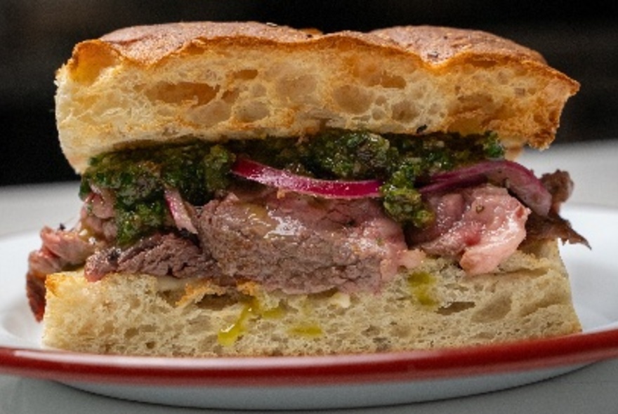 Close-up of a toasted focaccia sandwich, filled with rare beef and salsa verde, on a white plate. 