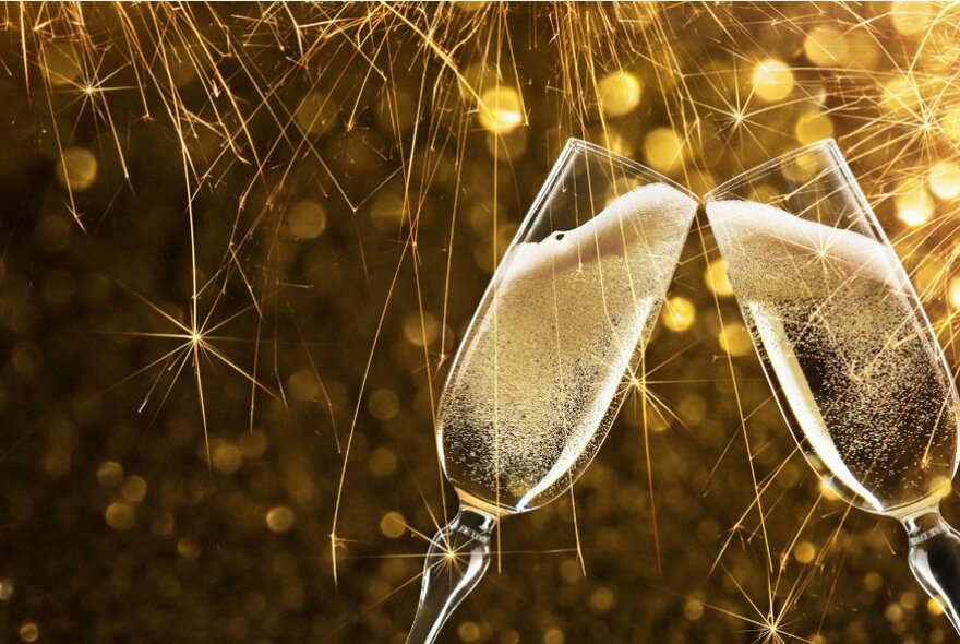 Two flute glass filled with sparkling wine leaning into each other, in front of a gold sparkly background.