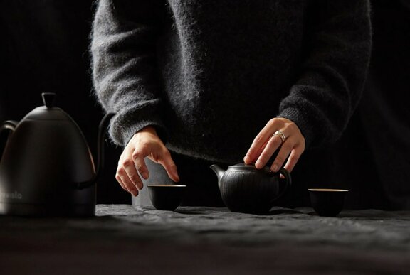 A Japanese teaset on a dark table, with a person behind wearing dark clothes, hands on the teacup.