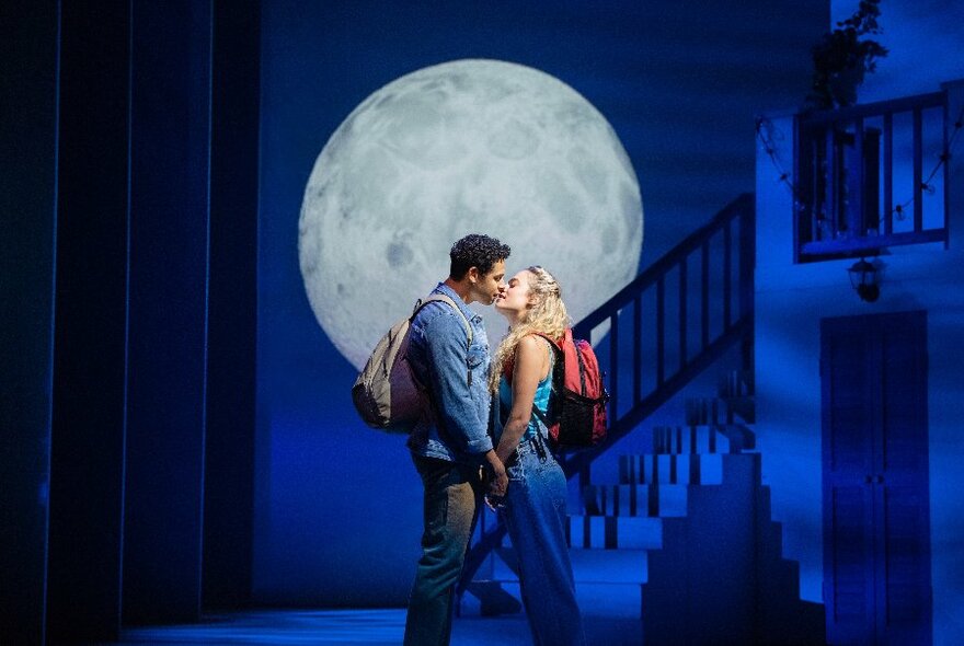 Two performers on a blue lit darkened theatre stage in front of a full moon prop, sharing a face-to-face kiss and holding hands.