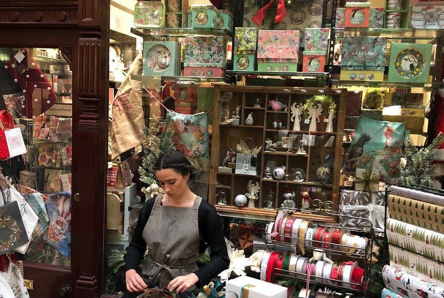 Woman wrapping a gift at the counter of store filled with stationery, ribbons and ornaments. 