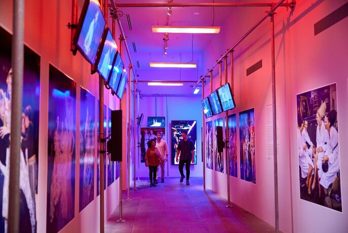 People walking through a hallway in a gallery with photos on the wall and neon lights.