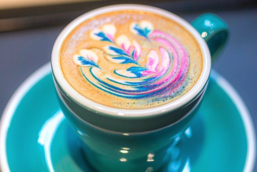 Coffee topped with pink and blue swirls in a turqoise cup and saucer.